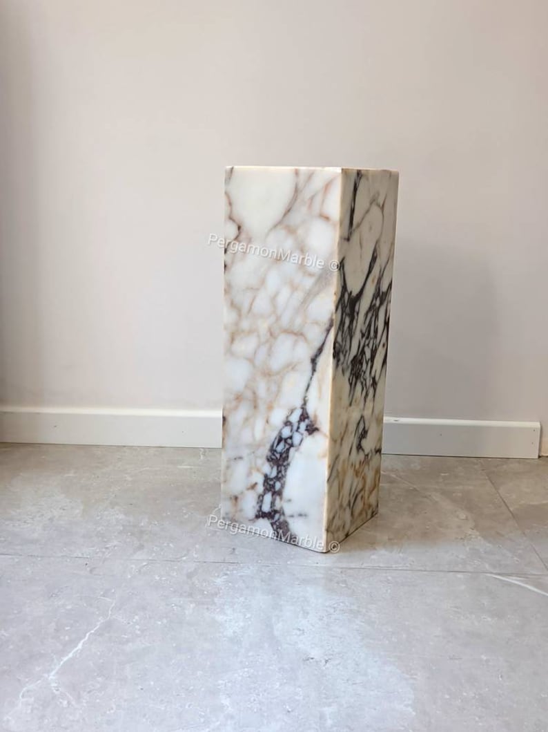 May include: A white marble pedestal with brown and gray veining. The pedestal is rectangular and has a square top. It is standing on a gray tiled floor.