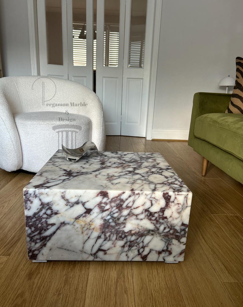 Op de afbeelding: Een vierkante salontafel van wit marmer met een draaiend patroon van paarse en bruine aderen. De tafel staat op een houten vloer.
