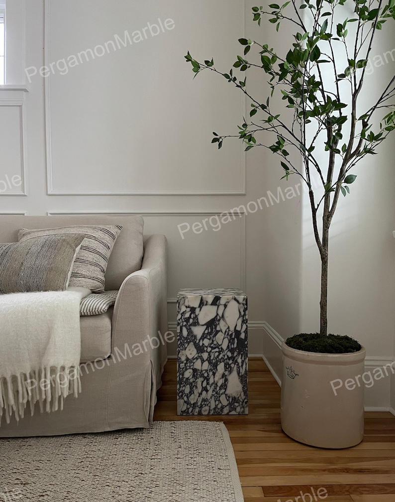 K&ouml;nnte beinhalten: Ein wei&szlig;es Sofa mit einer Wolldecke und zwei Kissen steht neben einem schwarzen und wei&szlig;en Marmor-Beistelltisch. Eine Topfpflanze mit gr&uuml;nen Bl&auml;ttern steht in der Ecke des Raumes.