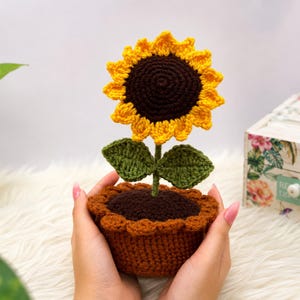 May include: A handcrafted crochet sunflower in a brown pot. The flower features a dark brown center with bright yellow petals and two green leaves on a stem. The pot has a textured brown base with a scalloped rim, all made from yarn.