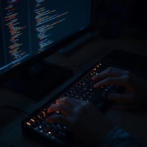 Puede incluir: Un monitor de computadora que muestra l&iacute;neas de c&oacute;digo, con un teclado retroiluminado en primer plano. La imagen muestra las manos de una persona escribiendo en el teclado. La pantalla muestra texto colorido, lo que sugiere desarrollo de software o programaci&oacute;n.