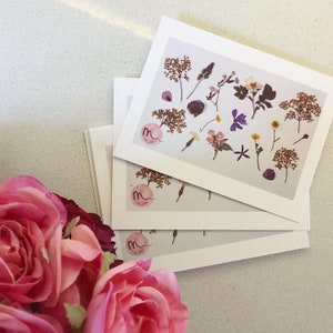 May include: A stack of white cards featuring pressed flowers in various colors and shapes. The cards have a pink logo with the letter "m". Pink roses are in the foreground.