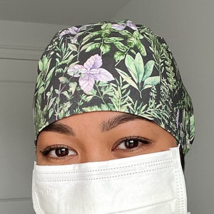 May include: A person wearing a black and green floral scrub cap with an adjustable strap. The person is also wearing a white face mask.