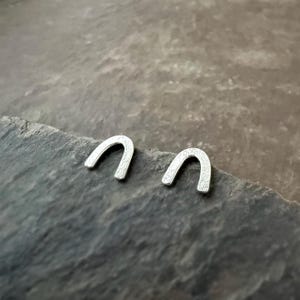 Op de afbeelding: Een paar kleine, zilverkleurige hoefijzer oorbellen. De oorbellen hebben een getextureerd oppervlak en worden gepresenteerd op een donker, getextureerd oppervlak. Eenvoudig, minimalistisch ontwerp.