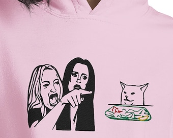 May include: A pink hoodie with a black and white image of the "Woman Yelling at Cat" meme. The meme features a woman yelling at a cat sitting across from her, with a plate of salad between them.