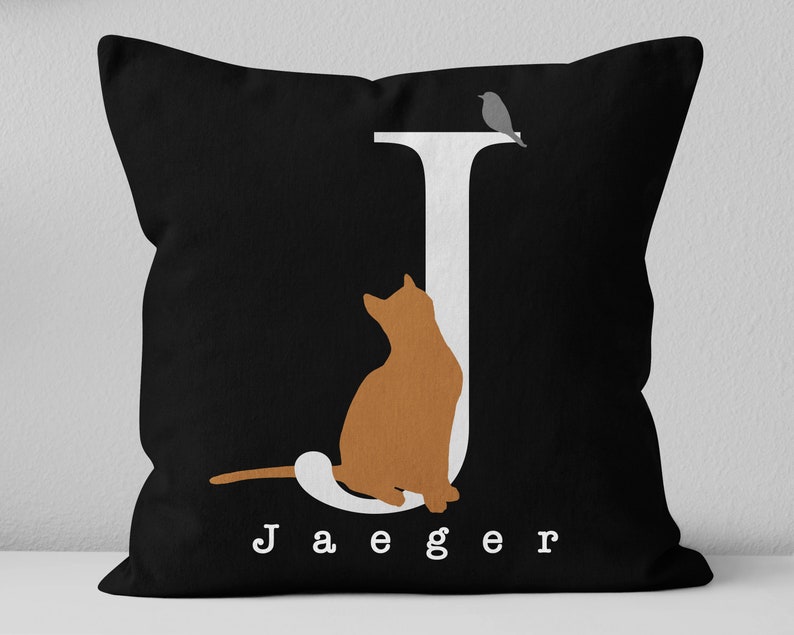 Puede incluir: Almohada negra con una letra J blanca, una silueta de gato y el nombre "Jaeger" impreso en blanco.