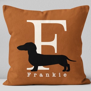 May include: A brown cushion with a white letter F and a black silhouette of a dachshund. The cushion has the name Frankie printed in white below the dog.