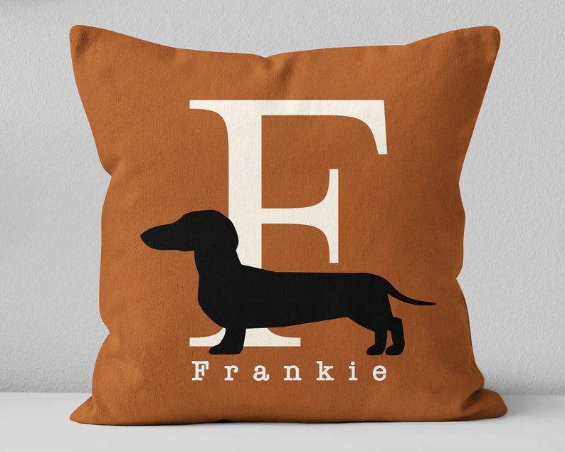 May include: A brown throw pillow with a white letter F and a black silhouette of a dachshund. The pillow has the name Frankie printed in white below the dog.