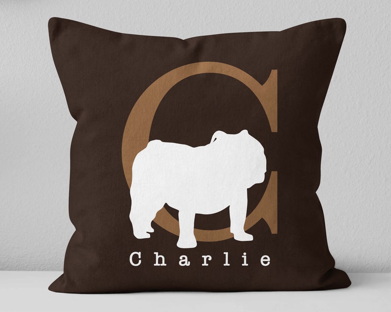 May include: Brown throw pillow with a white silhouette of a bulldog and the letter "C" in brown. The text "Charlie" is printed below the bulldog.