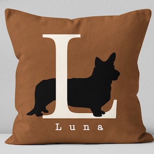 May include: A brown throw pillow with a white letter L and a black silhouette of a corgi dog. The dog is positioned to the right of the letter. The name "Luna" is printed below the letter.