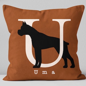 May include: A brown throw pillow with a white letter U and a black silhouette of a Boxer dog. The text "Uma" is printed below the letter.