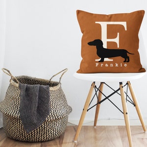 May include: A brown throw pillow with a white letter "F" and a black silhouette of a dachshund. The pillow has the name "Frankie" printed below the dachshund. The pillow is sitting on a white chair with wooden legs.