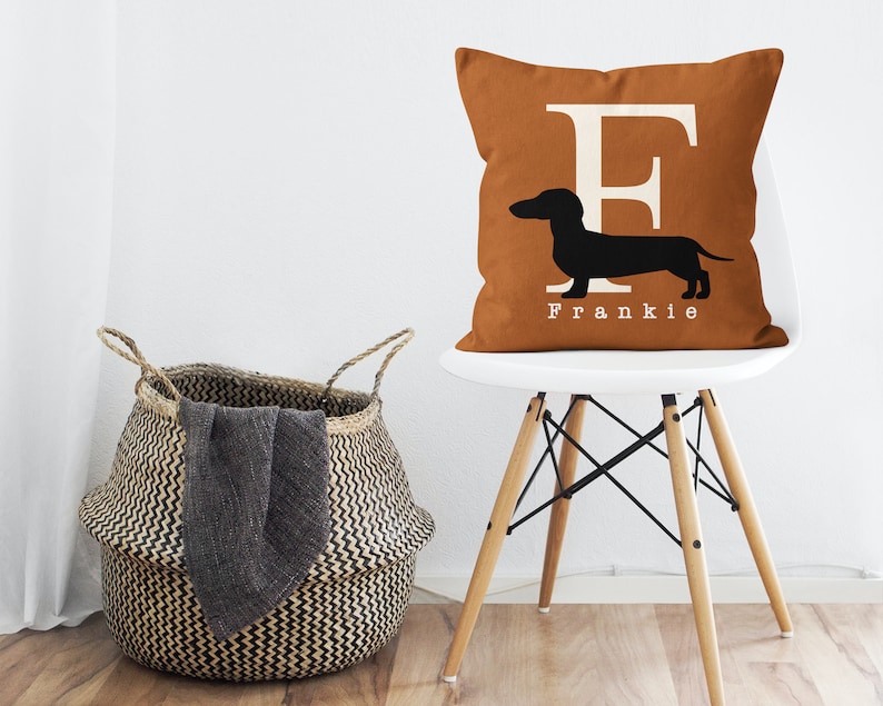 May include: A brown throw pillow with a white letter "F" and a black silhouette of a dachshund. The pillow has the name "Frankie" printed below the dachshund. The pillow is sitting on a white chair with wooden legs.