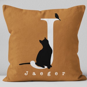 Puede incluir: Una almohada de color mostaza con una silueta blanca de un gato sentado junto a una letra J blanca. La almohada tiene el nombre "Jaeger" impreso en letras blancas debajo del gato.