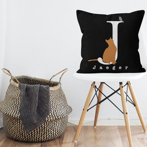 Puede incluir: Almohada negra y blanca con una letra J blanca, una silueta de gato y el nombre "Jaeger" impreso en la parte delantera. La almohada est&aacute; en una silla blanca con patas de madera.