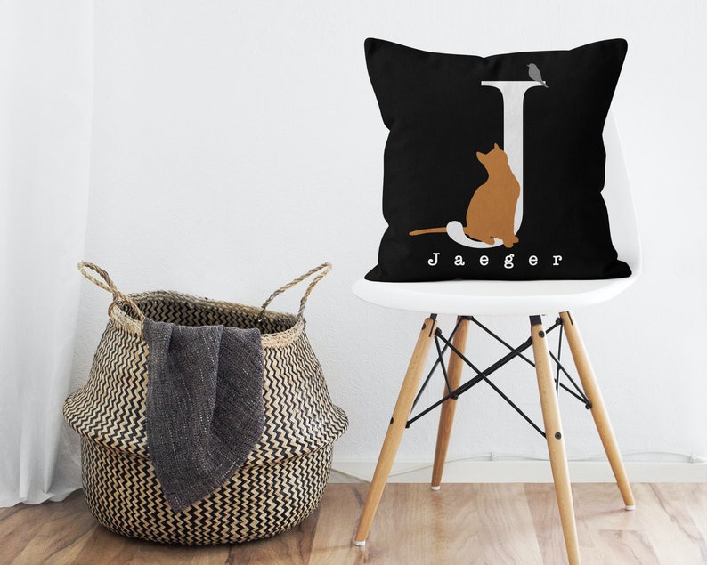 Puede incluir: Almohada negra y blanca con una letra J blanca, una silueta de gato y el nombre "Jaeger" impreso en la parte delantera. La almohada est&aacute; en una silla blanca con patas de madera.