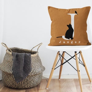Puede incluir: Una almohada de color mostaza con una letra J blanca, una silueta de gato negro y una peque&ntilde;a silueta de p&aacute;jaro. La almohada tiene el nombre "Jaeger" impreso en blanco debajo de la letra J.