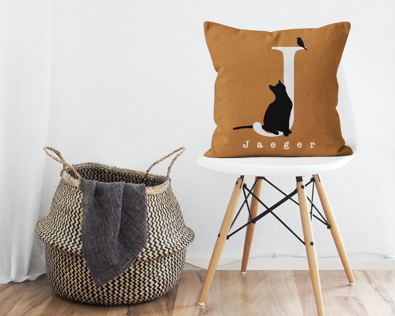 Puede incluir: Una almohada de color mostaza con una letra J blanca, una silueta de gato negro y una peque&ntilde;a silueta de p&aacute;jaro. La almohada tiene el nombre "Jaeger" impreso en blanco debajo de la letra J.