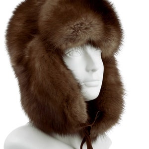 Puede incluir: Un gorro de piel marrón con orejeras y una corbata de ante marrón. El gorro está en una cabeza de maniquí blanca.