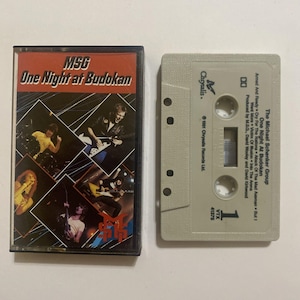 May include: A cassette tape and its plastic case for The Michael Schenker Group album One Night at Budokan. The case insert features live performance photos and the text MSG One Night at Budokan. The beige cassette shell lists track titles.
