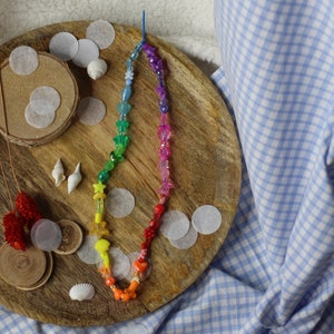 Puede incluir: Un collar de cuentas de colores arcoíris con varios encantos, incluyendo estrellas, corazones y conchas marinas. El collar está colocado en una bandeja de madera con círculos de papel blanco y otros artículos decorativos.