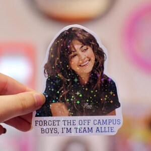 Pode incluir: Um adesivo recortado com uma pessoa sorridente de cabelos castanhos longos e ondulados vestindo uma camiseta preta. O adesivo tem um efeito hologr&aacute;fico de estrelas e o texto "FORGET THE OFF CAMPUS BOYS, I'M TEAM ALLIE." em letras azuis mai&uacute;sculas.