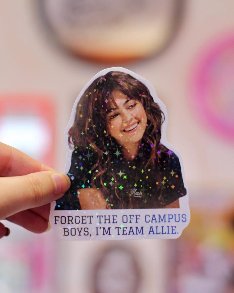 Pode incluir: Um adesivo recortado com uma pessoa sorridente de cabelos castanhos longos e ondulados vestindo uma camiseta preta. O adesivo tem um efeito hologr&aacute;fico de estrelas e o texto "FORGET THE OFF CAMPUS BOYS, I'M TEAM ALLIE." em letras azuis mai&uacute;sculas.