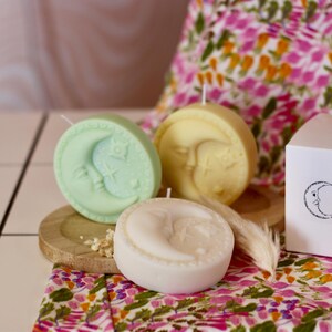 May include: Three round candles in shades of green, yellow, and white, each featuring a crescent moon and star design. The candles sit on a wooden plate, with a floral patterned fabric in the background.