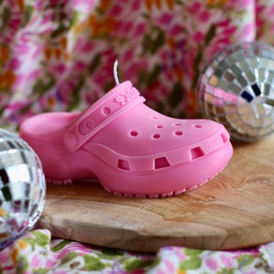 Puede incluir: Una vela de zueco de goma rosa con la palabra "Crocs" en relieve en la correa. La vela está sobre una superficie de madera con una bola de discoteca en el fondo.