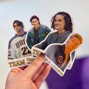 Pode incluir: Quatro autocolantes hologr&aacute;ficos com retratos de pessoas. Um autocolante mostra uma pessoa com uma camisola desportiva e o texto TEAM. Outro autocolante mostra uma pessoa com um casaco escuro e o texto TEAM DI LAURENTIS. Acabamento hologr&aacute;fico com estrelas.