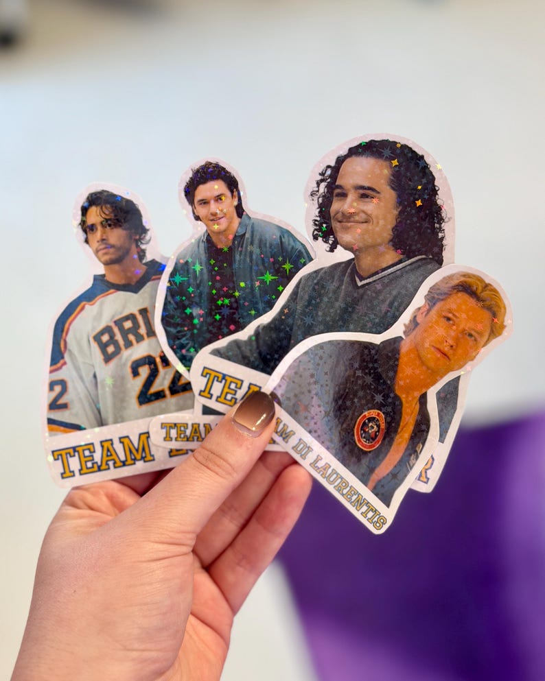 Pode incluir: Quatro autocolantes hologr&aacute;ficos com retratos de pessoas. Um autocolante mostra uma pessoa com uma camisola desportiva e o texto TEAM. Outro autocolante mostra uma pessoa com um casaco escuro e o texto TEAM DI LAURENTIS. Acabamento hologr&aacute;fico com estrelas.