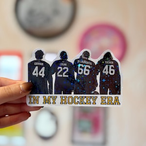 Pode incluir: Um adesivo hologr&aacute;fico com quatro jogadores de h&oacute;quei vistos por tr&aacute;s, vestindo camisas com os nomes Graham, Logan, Di Laurentis e Tucker, e os n&uacute;meros 44, 22, 66 e 46. O texto IN MY HOCKEY ERA est&aacute; impresso em amarelo na parte inferior.