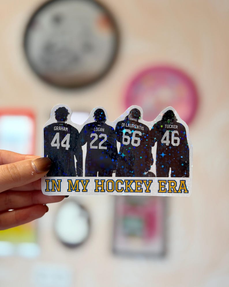 Pode incluir: Um adesivo hologr&aacute;fico com quatro jogadores de h&oacute;quei vistos por tr&aacute;s, vestindo camisas com os nomes Graham, Logan, Di Laurentis e Tucker, e os n&uacute;meros 44, 22, 66 e 46. O texto IN MY HOCKEY ERA est&aacute; impresso em amarelo na parte inferior.