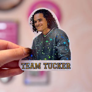 Pode incluir: Um adesivo hologr&aacute;fico com uma pessoa de cabelos escuros cacheados vestindo um su&eacute;ter cinza com decote em V. O adesivo tem um acabamento com padr&atilde;o de estrelas e inclui o texto "TEAM TUCKER" em letras azuis em negrito com contorno amarelo.