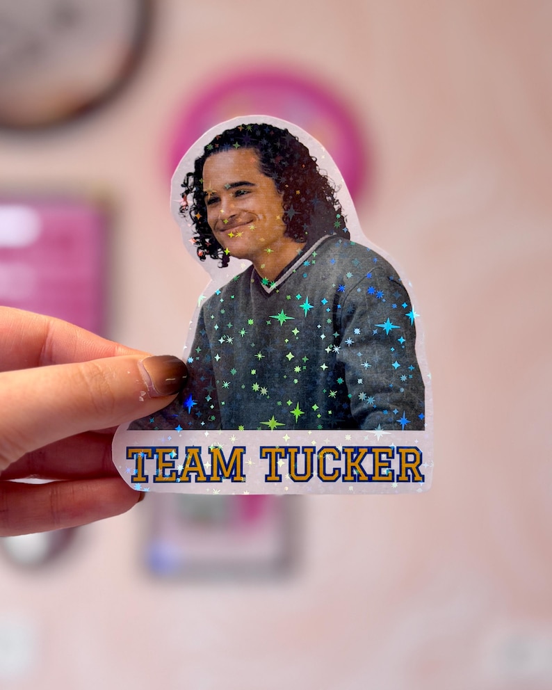 Pode incluir: Um adesivo hologr&aacute;fico com uma pessoa de cabelos escuros cacheados vestindo um su&eacute;ter cinza com decote em V. O adesivo tem um acabamento com padr&atilde;o de estrelas e inclui o texto "TEAM TUCKER" em letras azuis em negrito com contorno amarelo.