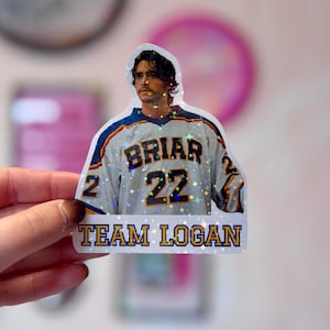 Pode incluir: Um autocolante hologr&aacute;fico com uma pessoa de cabelo escuro a usar uma camisola de h&oacute;quei branca, azul e amarela com o texto BRIAR e o n&uacute;mero 22. Por baixo da camisola, o texto TEAM LOGAN est&aacute; impresso em letras azuis e amarelas.