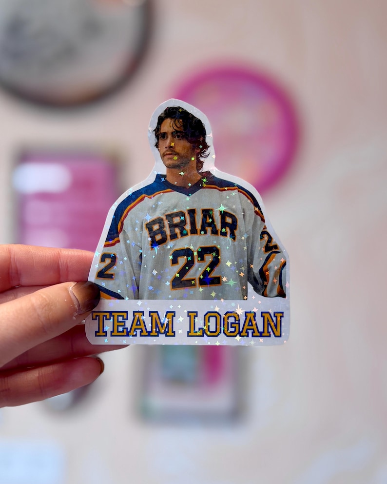 Pode incluir: Um autocolante hologr&aacute;fico com uma pessoa de cabelo escuro a usar uma camisola de h&oacute;quei branca, azul e amarela com o texto BRIAR e o n&uacute;mero 22. Por baixo da camisola, o texto TEAM LOGAN est&aacute; impresso em letras azuis e amarelas.