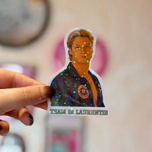 Pode incluir: Um autocolante hologr&aacute;fico recortado com o retrato de uma pessoa de cabelo loiro a usar um casaco escuro com um emblema circular no peito. O autocolante tem um efeito de brilho em forma de estrela e o texto "TEAM DI LAURENTIS" em letras amarelas.