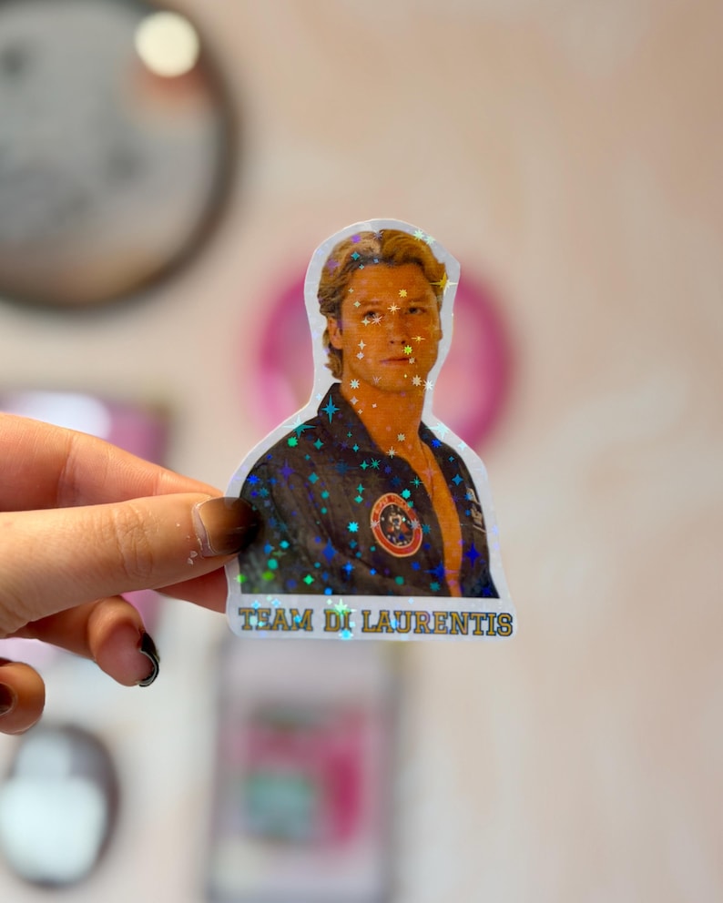 Pode incluir: Um autocolante hologr&aacute;fico recortado com o retrato de uma pessoa de cabelo loiro a usar um casaco escuro com um emblema circular no peito. O autocolante tem um efeito de brilho em forma de estrela e o texto "TEAM DI LAURENTIS" em letras amarelas.