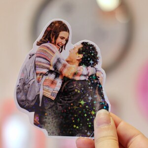 Pode incluir: Um autocolante hologr&aacute;fico recortado que mostra um casal a abra&ccedil;ar-se. A pessoa &agrave; esquerda usa uma camisola &agrave;s riscas multicolor e uma mochila cinzenta, enquanto a pessoa &agrave; direita usa um casaco escuro. O autocolante tem um acabamento hologr&aacute;fico de estrelas.