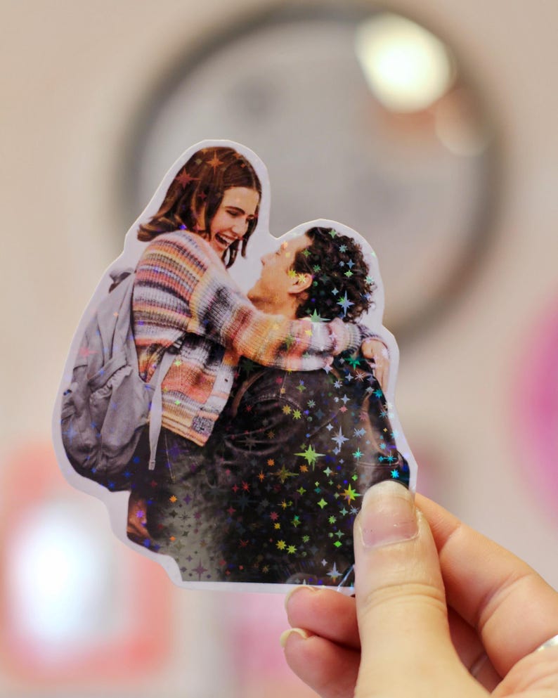 Pode incluir: Um autocolante hologr&aacute;fico recortado que mostra um casal a abra&ccedil;ar-se. A pessoa &agrave; esquerda usa uma camisola &agrave;s riscas multicolor e uma mochila cinzenta, enquanto a pessoa &agrave; direita usa um casaco escuro. O autocolante tem um acabamento hologr&aacute;fico de estrelas.