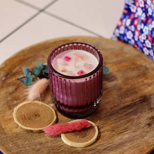 Puede incluir: Un portavelas de cristal morado con una vela blanca, decorada con flores rosas y cristales. La vela se encuentra sobre una bandeja de madera con rodajas de madera y detalles decorativos rosas. El portavelas tiene un diseño acanalado.