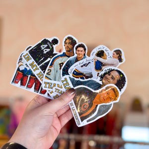 Pode incluir: Uma cole&ccedil;&atilde;o de adesivos hologr&aacute;ficos com v&aacute;rias cenas e personagens de uma s&eacute;rie de televis&atilde;o sobre h&oacute;quei. Os adesivos incluem imagens de jogadores com camisas, um casal se abra&ccedil;ando e o texto IN MY HO, TEAM e LAURENTIS.