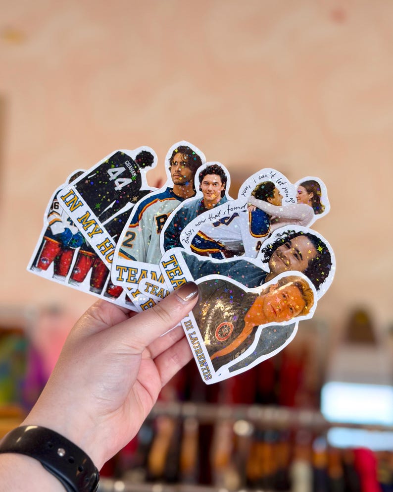 Pode incluir: Uma cole&ccedil;&atilde;o de adesivos hologr&aacute;ficos com v&aacute;rias cenas e personagens de uma s&eacute;rie de televis&atilde;o sobre h&oacute;quei. Os adesivos incluem imagens de jogadores com camisas, um casal se abra&ccedil;ando e o texto IN MY HO, TEAM e LAURENTIS.