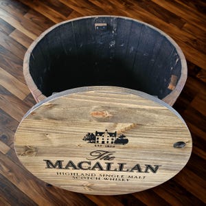 May include: Wooden barrel with a lid, featuring the text "The Macallan Highland Single Malt Scotch Whisky." The lid displays a black graphic of a building and the text "EST 1824." The barrel is made of light brown wood with a dark interior.