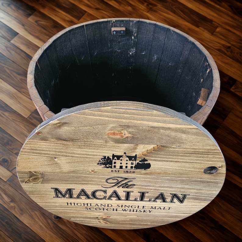 May include: Wooden barrel with a lid, featuring the text "The Macallan Highland Single Malt Scotch Whisky." The lid displays a black graphic of a building and the text "EST 1824." The barrel is made of light brown wood with a dark interior.