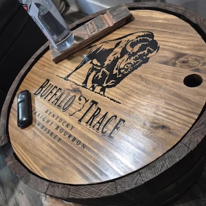 May include: A wooden barrel lid with the words "Buffalo Trace Kentucky Straight Bourbon Whiskey" and a black bison graphic. A black lighter and a bottle rest on the lid. The lid is made of light brown wood.
