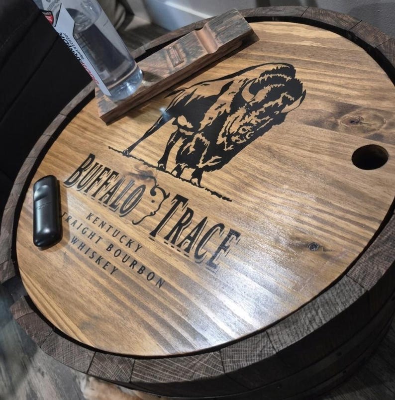 May include: A wooden barrel lid with the words "Buffalo Trace Kentucky Straight Bourbon Whiskey" and a black bison graphic. A black lighter and a bottle rest on the lid. The lid is made of light brown wood.