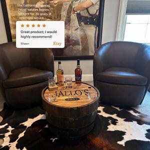 May include: A wooden barrel table with a round top, featuring the text "JALLO'S EST. 2022". Two brown leather armchairs flank the table. The table holds two bottles of liquor and two glasses. A black and white cowhide rug is on the floor.