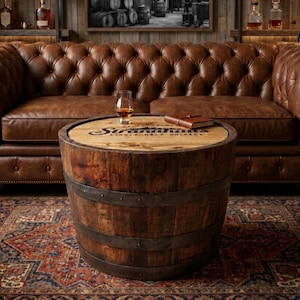 May include: A round coffee table crafted from a repurposed wooden barrel with dark metal bands. The tabletop features the text Siranahan Single Malt Whiskey, a glass of amber liquid, and a small brown leather-bound notebook resting on the surface.